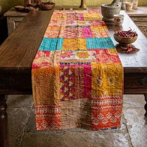 May include: A colorful patchwork table runner with a variety of patterns and colors, including orange, yellow, pink, and blue. The runner is placed on a dark wooden table. A brass kitchen tool is in the background.