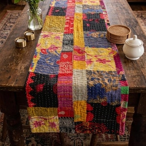May include: A colorful patchwork table runner with a quilted design. The runner features a variety of patterns and colors, including red, yellow, blue, and black. It is placed on a rustic wooden table, with a vase of flowers and other decorative items.