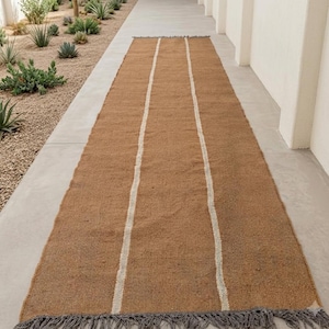 May include: A long, rectangular runner rug featuring a textured, tan-colored woven surface. The rug is accented by two vertical, off-white stripes running the length of the piece and finished with dark gray fringe along the short ends.
