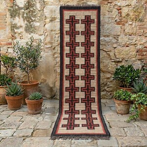 May include: A long, rectangular rug with a repeating cross pattern in black and red on a beige background. The rug has a dark border and fringe. The rug is displayed outdoors, leaning against a stone wall, with potted plants nearby.