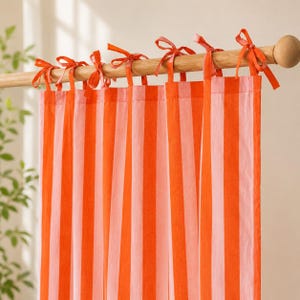 May include: A curtain featuring vertical stripes in alternating shades of bright orange and soft pink. The fabric is suspended from a light-colored wooden rod by several fabric ties knotted into bows along the top edge of the panel.