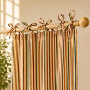 Op de afbeelding: Een gordijn met verticale strepen in de kleuren oranje, geel, donkergroen en wit. De stof hangt aan een lichtgekleurde houten roede door middel van diverse stoffen linten die boven aan de gordijnkop tot strikken zijn geknoopt.