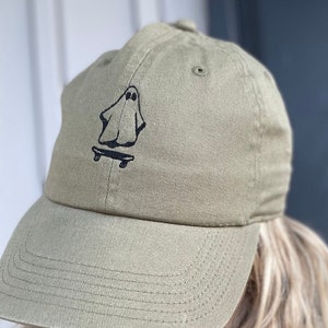 May include: Olive green baseball cap featuring a black graphic of a ghost riding a skateboard. The cap has a curved brim and a fabric strap closure. The cap is made of a durable, textured fabric.