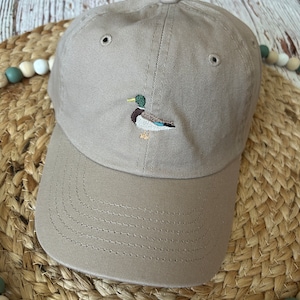 May include: A tan baseball cap with an embroidered duck design. The duck has green, blue, and brown details. The hat has a curved brim and a classic six-panel design. The hat is made of a soft, durable fabric.