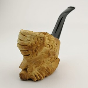 May include: A carved wooden pipe with a black stem. The bowl of the pipe is shaped like a face with intricate details.