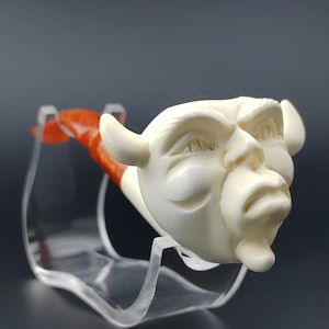 May include: A white ceramic pipe with a carved face and horns. The pipe is on a clear plastic stand. The pipe is a unique and interesting piece of art.