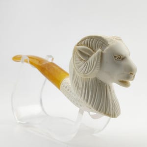 May include: A carved ivory-colored pipe featuring a ram's head design. The pipe has a yellow-orange stem and a decorative white band. The ram's head is detailed with carved fur and horns. The pipe is displayed on a clear stand.