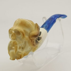 Puede incluir: Una pipa de calavera blanca y azul con un diseño de espiral azul y blanco en el tallo. La pipa está en un soporte de acrílico transparente.