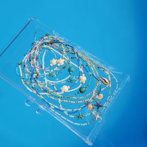 Könnte beinhalten: Eine Sammlung von Perlenketten mit kleinen, bunten Glasperlen in Blau-, Weiß- und Rosatönen. Die Ketten sind mit Anhängern in Form von weißen und türkisfarbenen Meeresschildkröten, Seesternen und Muscheln verziert und mit Metallverschlüssen versehen.