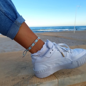 Könnte beinhalten: Fußkettchen mit geometrischem Muster in Blau-, Braun- und Weißtönen. Das Armband wird mit weißen Turnschuhen und einer blauen Jeans getragen. Der Hintergrund zeigt einen Strand und das Meer.