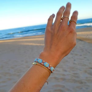 Könnte beinhalten: Ein gewebtes Freundschaftsarmband mit Rautenmuster in hellblauen, weißen und braunen Fäden, kombiniert mit einem dünnen Armband aus goldfarbenem Metall. Zwei schlichte Ringe aus goldfarbenem Metall werden am Zeige- und Mittelfinger getragen.