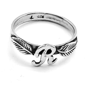 Peut inclure: Une bague en argent avec une lettre "R" stylisée et deux motifs de feuilles de chaque côté. La bague est marquée "925" et d'un numéro "4" à l'intérieur.