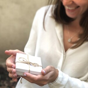Peut inclure: Une bo&icirc;te cadeau blanche attach&eacute;e avec de la ficelle, tenue par une personne portant une chemise blanche. La personne sourit et regarde la bo&icirc;te.