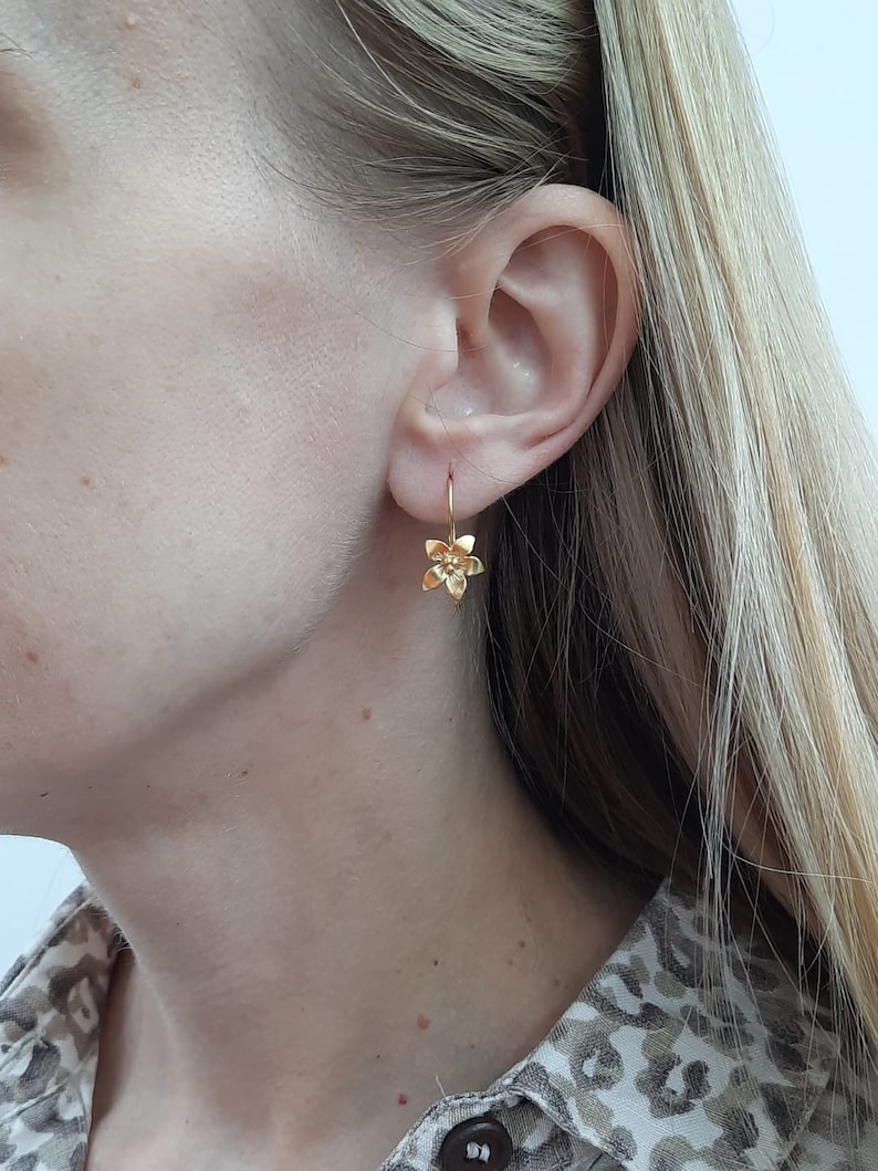 Peut inclure: Boucles d'oreilles en forme de fleur dor&eacute;es avec un design d&eacute;licat. Les boucles d'oreilles pendent &agrave; l'oreille d'une personne.