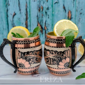 May include: Two copper mugs with black handles and intricate floral designs. The mugs are filled with a clear liquid and garnished with lemon slices and mint leaves. The mugs are sitting on a white marble surface with a book that says 'ERIZA'.