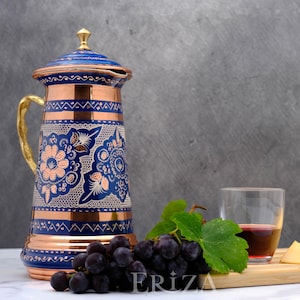 May include: A blue and copper jug with intricate floral designs and a gold handle. The jug is sitting on a white surface with a bunch of grapes and a glass of red wine.