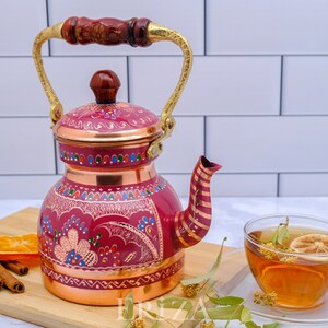 May include: A red copper teapot with a gold handle and a floral design. The teapot is sitting on a wooden cutting board with a cup of tea and a lemon slice.