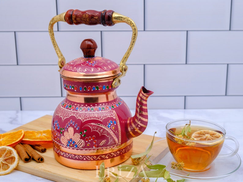 May include: A red copper teapot with a gold handle and a floral design. The teapot is sitting on a wooden cutting board with a cup of tea and a lemon slice.