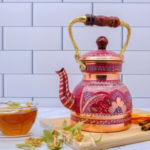 May include: A red copper teapot with a gold handle and intricate painted designs. The teapot is sitting on a wooden cutting board with cinnamon sticks, dried orange slices, and a cup of tea.