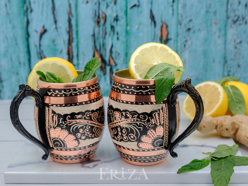 May include: Two copper mugs with black handles and intricate floral designs. The mugs are filled with a clear liquid and garnished with lemon slices and mint leaves. The mugs are sitting on a white marble surface with a book that says 'ERIZA'.