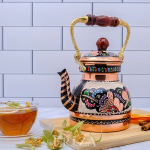 May include: A copper tea kettle with a wooden handle and a colorful floral design. The kettle is sitting on a wooden cutting board with cinnamon sticks, orange slices, and a cup of tea.
