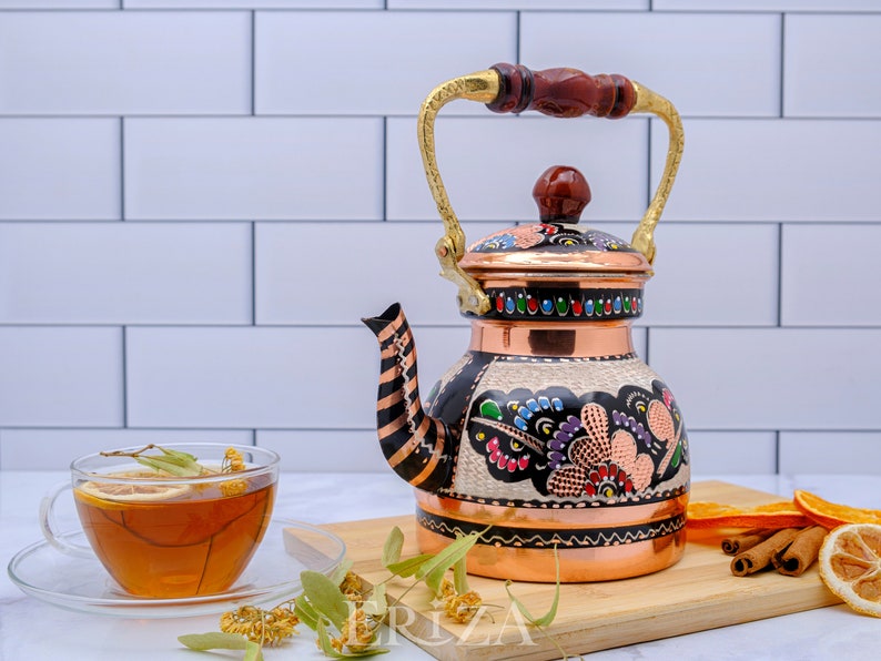 May include: A copper tea kettle with a wooden handle and a colorful floral design. The kettle is sitting on a wooden cutting board with cinnamon sticks, orange slices, and a cup of tea.