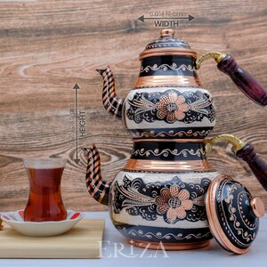 Puede incluir: Una tetera de cobre de dos niveles con dise&ntilde;os florales en blanco y negro y detalles en cobre. La tetera tiene dos asas de madera y una tapa. La tetera mide 31,1 cm de alto y 16,8 cm de ancho.