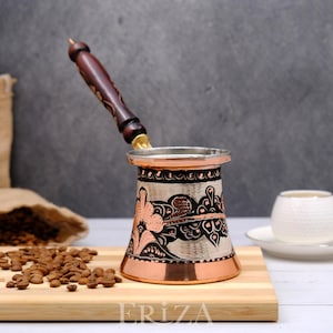 Puede incluir: Una cafetera turca tradicional con cuerpo de cobre y mango de madera. La cafetera está decorada con intrincados patrones florales en negro y cobre. La cafetera está sobre una tabla de cortar de madera con granos de café esparcidos a su alrededor.