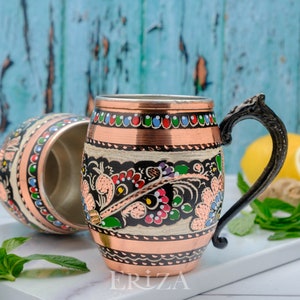 May include: Two copper mugs with black and white floral designs and colorful accents. The mugs have a rounded shape and a handle. The mug on the right has the word "ERIZA" printed on the bottom.