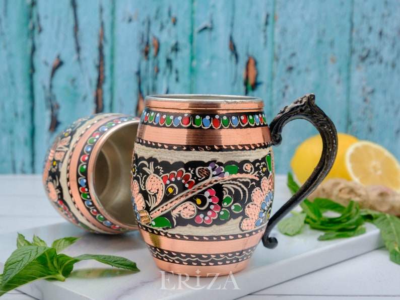May include: Two copper mugs with black and white floral designs and colorful accents. The mugs have a rounded shape and a handle. The mug on the right has the word "ERIZA" printed on the bottom.