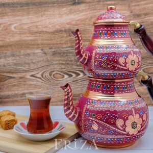 Puede incluir: Un juego de t&eacute; turco tradicional con una tetera de cobre de dos niveles decorada con coloridos patrones florales. La tetera tiene un asa de madera y un vaso de t&eacute; est&aacute; en primer plano.