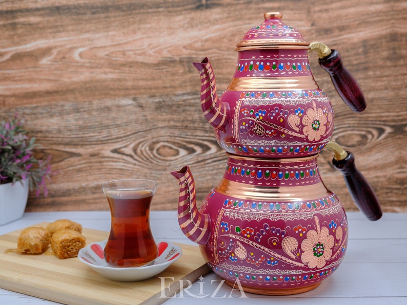 Puede incluir: Un juego de t&eacute; turco tradicional con una tetera de cobre de dos niveles decorada con coloridos patrones florales. La tetera tiene un asa de madera y un vaso de t&eacute; est&aacute; en primer plano.