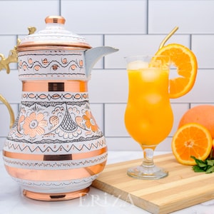 May include: White and copper pitcher with floral designs and a gold handle. The pitcher is on a wooden surface with a glass of orange juice and orange slices.