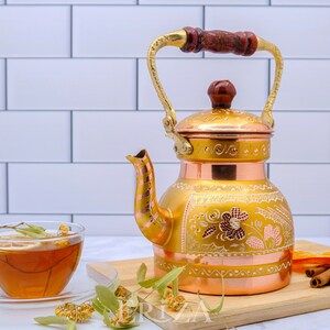 May include: A gold and copper tea kettle with intricate floral designs and a wooden handle. The kettle is sitting on a wooden cutting board with a cup of tea, dried flowers, and cinnamon sticks.