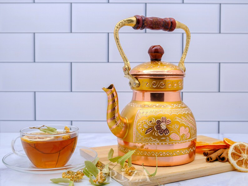 May include: A gold and copper tea kettle with intricate floral designs and a wooden handle. The kettle is sitting on a wooden cutting board with a cup of tea, dried flowers, and cinnamon sticks.