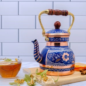 May include: A blue and copper tea kettle with a floral design. The kettle has a wooden handle and a brass spout. The kettle is sitting on a wooden cutting board with cinnamon sticks, orange slices, and dried flowers.