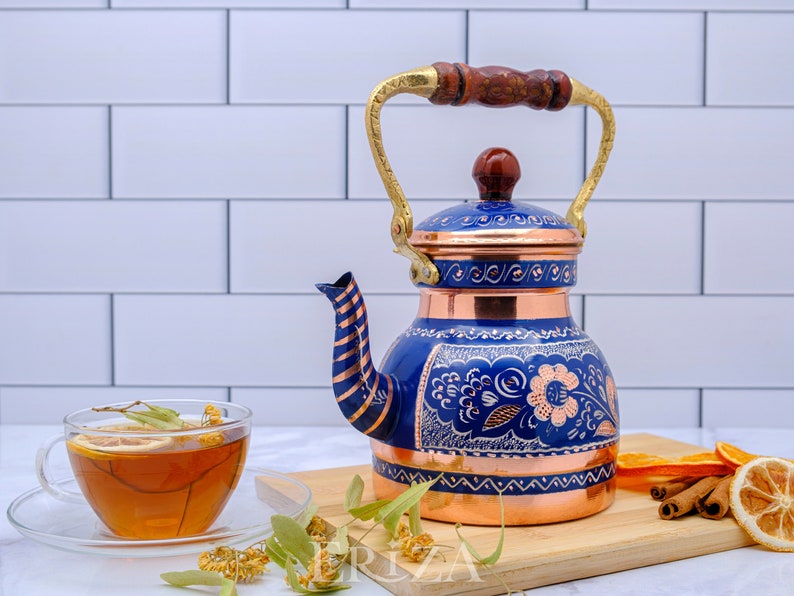 May include: A blue and copper tea kettle with a floral design. The kettle has a wooden handle and a brass spout. The kettle is sitting on a wooden cutting board with cinnamon sticks, orange slices, and dried flowers.