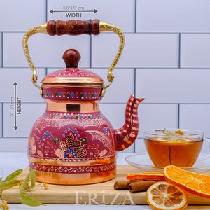 May include: A red copper teapot with a gold handle and intricate floral designs. The teapot is 9.1 inches tall and 6.8 inches wide. It is sitting on a wooden surface with cinnamon sticks and orange slices.