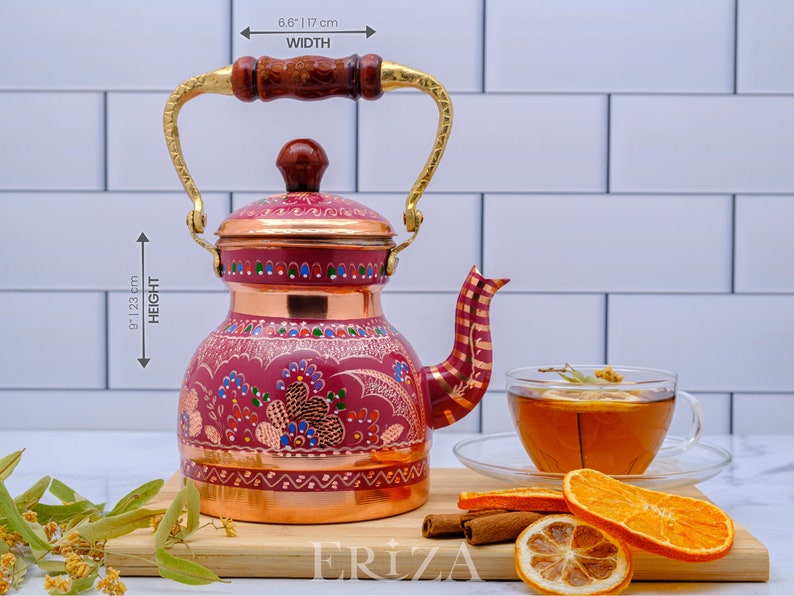 May include: A red copper teapot with a gold handle and intricate floral designs. The teapot is 9.1 inches tall and 6.8 inches wide. It is sitting on a wooden surface with cinnamon sticks and orange slices.
