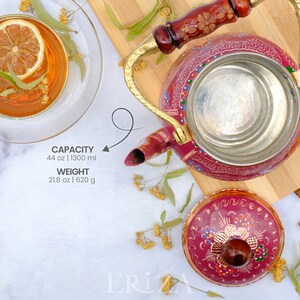 May include: A pink metal teapot with a gold handle and lid. The teapot has a floral design painted on it. The teapot is sitting on a wooden cutting board. The teapot has a capacity of 44 oz | 1300 ml and weighs 21.8 oz | 620 g.
