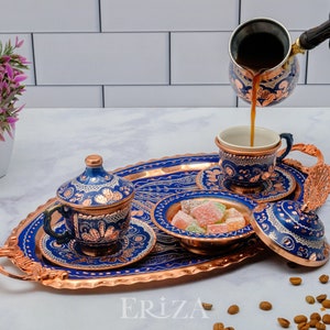May include: A copper serving tray with blue and gold enamel designs. The tray holds a set of Turkish coffee cups, a sugar bowl, and a coffee pot. The coffee pot is pouring coffee into one of the cups. The tray is surrounded by coffee beans.