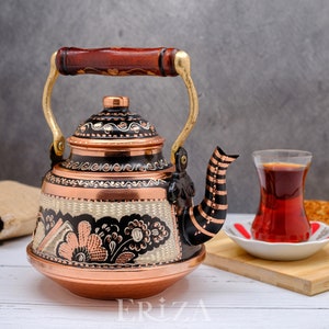 May include: A traditional Turkish copper teapot with a black and copper floral design and a wooden handle. The teapot is filled with tea and is sitting on a white wooden table. A glass of tea and a plate of Turkish bread are also on the table.
