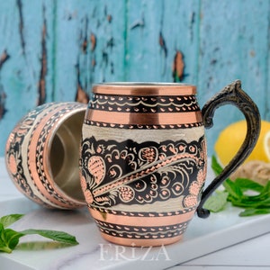 May include: A copper mug with a black and white floral design and a copper handle. The mug is sitting on a white surface with a sprig of mint and a lemon.