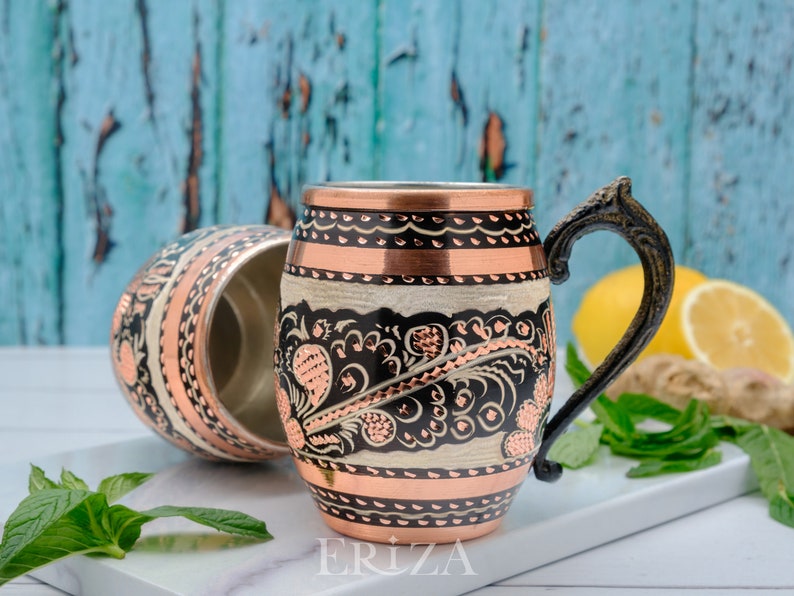 May include: A copper mug with a black and white floral design and a copper handle. The mug is sitting on a white surface with a sprig of mint and a lemon.