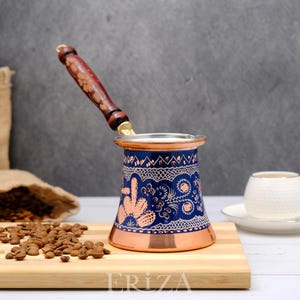 Pode incluir: Uma cafeteira turca tradicional com um design azul e cobre. A cafeteira tem uma pega de madeira e é decorada com padrões florais intrincados. A cafeteira está sentada sobre uma superfície de madeira com grãos de café espalhados à sua volta.