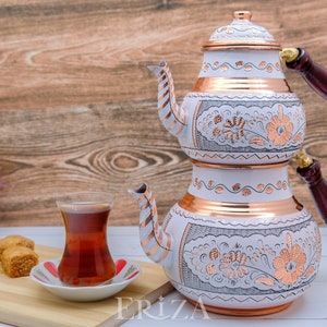 Puede incluir: Un juego de t&eacute; turco blanco y cobre con dise&ntilde;os florales. La tetera tiene dos niveles y un mango de madera. Un vaso de t&eacute; y un plato de baklava est&aacute;n sobre una tabla de cortar de madera.
