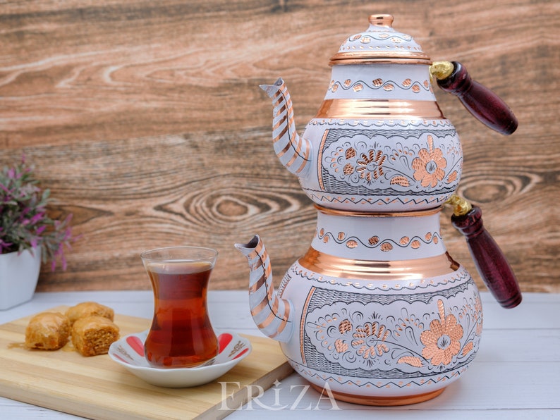 Puede incluir: Un juego de t&eacute; turco blanco y cobre con dise&ntilde;os florales. La tetera tiene dos niveles y un mango de madera. Un vaso de t&eacute; y un plato de baklava est&aacute;n sobre una tabla de cortar de madera.