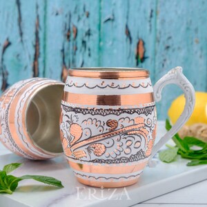 May include: Two white and copper mugs with intricate floral designs. The mugs have a white handle and are sitting on a white surface.