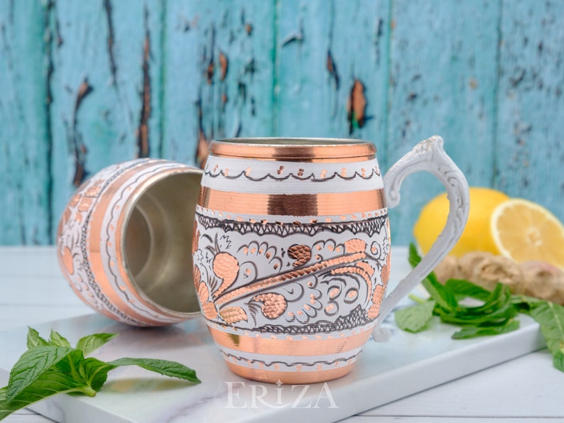 May include: Two white and copper mugs with intricate floral designs. The mugs have a white handle and are sitting on a white surface.