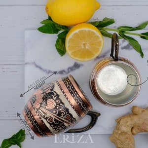 May include: Two copper mugs with intricate designs. The mug on the left has a black and white floral pattern with copper accents. The mug on the right is plain copper. Both mugs have a capacity of 18.5 ounces or 550 milliliters and weigh 7.23 ounces or 205 grams.
